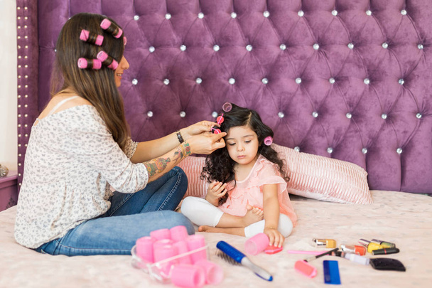 Mother curling hair of daughter with curlers while sitting on bed - Photo, Image