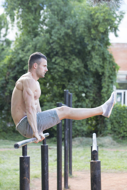 Jovem musculoso com um corpo perfeito fazendo exercícios nas barras paralelas no parque
 - Foto, Imagem