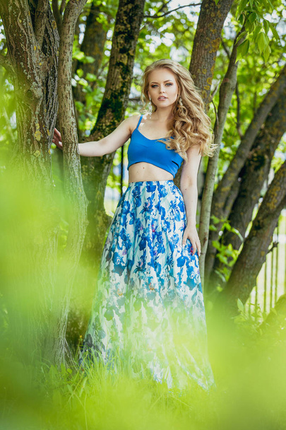 Beautiful young girl in a summer dress at sunset. Fashion photo in the forest. Model in a blue top and long skirt, with flowing curly hair. - Фото, зображення
