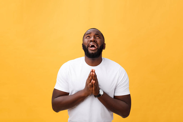 Studio portrait of young African American man in white shirt, holding hands in prayer, looking at the camera with thoughtful skeptical expression on his face, suspecting of something. Body language. - Photo, Image