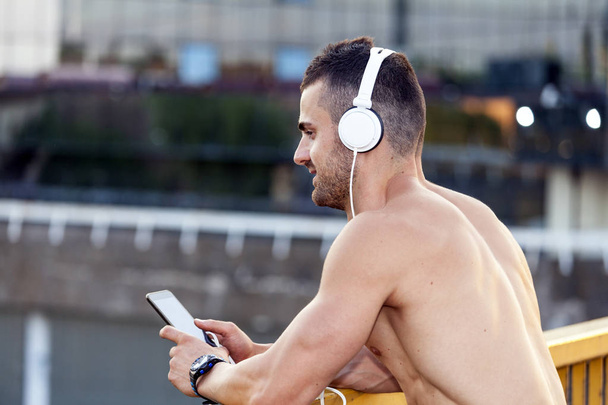 Handsome and attractive muscular young man, shirtless resting on the bridge. He listens to music and looks around. He has an excellent view of the river and the city - Photo, Image