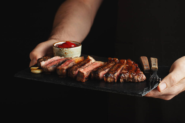 Un homme tenant un steak de bœuf grillé juteux avec des épices et de la sauce rouge sur une planche à découper en pierre sur fond noir
 - Photo, image