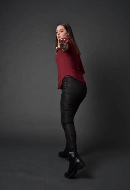 赤いシャツと革のズボンを身に着けているブルネットの女の子の完全な長さの肖像画。灰色のスタジオの背景上の立ちポーズ. - 写真・画像