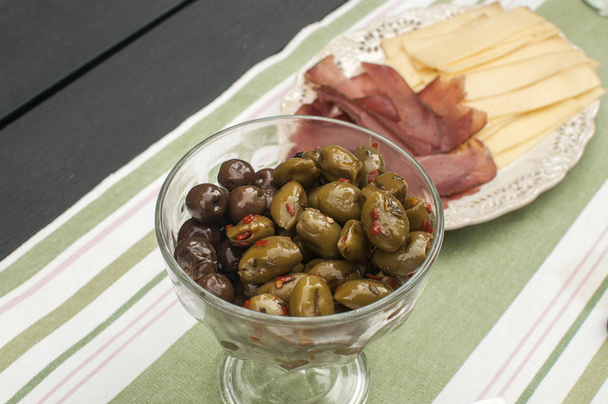 Скляна миска з оливками в оливковій олії та блюдце з пастрамі та сиром як закуски крупним планом на покритому текстильним килимком домашнього столу
 - Фото, зображення