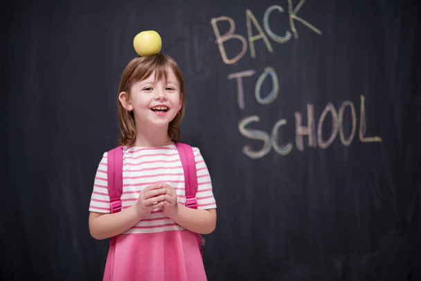 happy child holding apple on head with back to shool drawing on chalkboard in background - Photo, Image