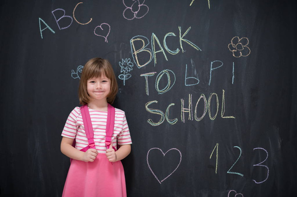 Happy school girl child with backpack writing  back to school on black chalkboard - 写真・画像