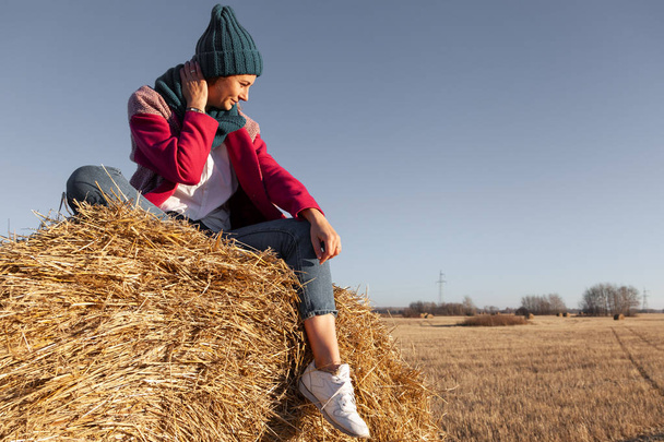 A young woman in a blue knitted hat, pink coat and jeans  enjoys nature, sitting on a haystack around blue sky. The concept of livestyle and outdoor recreation in autumn - Фото, изображение