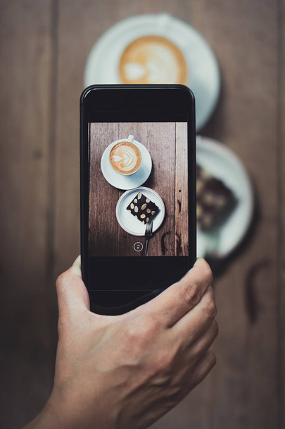 Top view of Hand holding mobile take a photo of coffee cup and cake on wood table in cafe restaurant.leisure lifestyle - Photo, image
