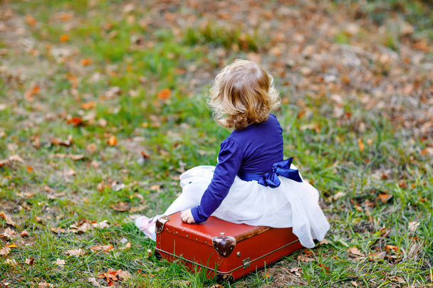 Cute little toddler girl sitting on suitcase in autumn park. Happy healthy baby enjoying walking with parents. Sunny warm fall day with child. Active leisure and activity with kids in nature. - 写真・画像
