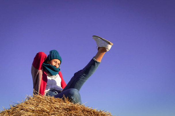 Portrait of a beautiful young model in  blue knitted hat  and warm clothes  have fun,  sitting on haystacks in  sunny autumn day . Autumn warm photo. Woman smiling and look away, joyful cheerful mood. - Photo, Image