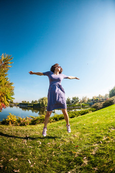 A girl in a dress runs through a green meadow. Woman walking in the park. Activity in nature. Slender brunette is having fun. Girl jumping on the grass. - Photo, image