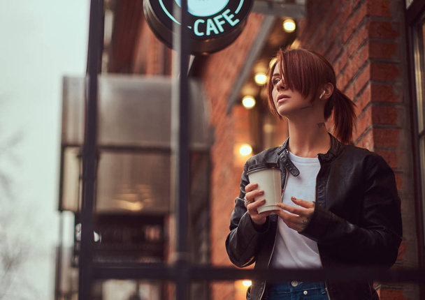 Urocza dziewczyna na sobie skórzaną kurtkę z plecakiem trzymając kubek z kawa na wynos poza w pobliżu kawiarni. - Zdjęcie, obraz