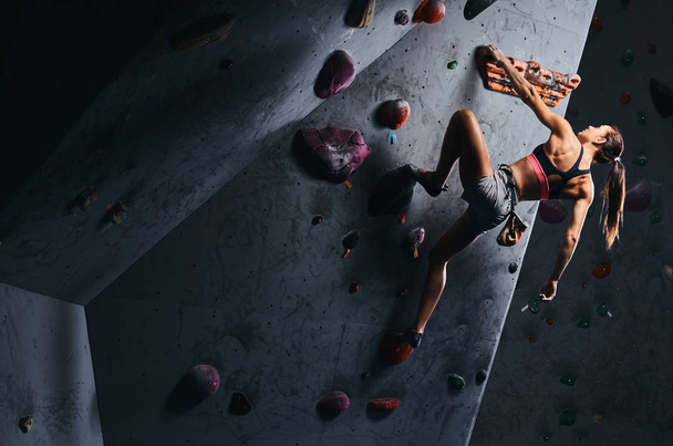 Νεαρή γυναίκα στο σορτσάκι και το αθλητικό σουτιέν άσκηση σε έναν τοίχο bouldering σε εσωτερικούς χώρους. - Φωτογραφία, εικόνα