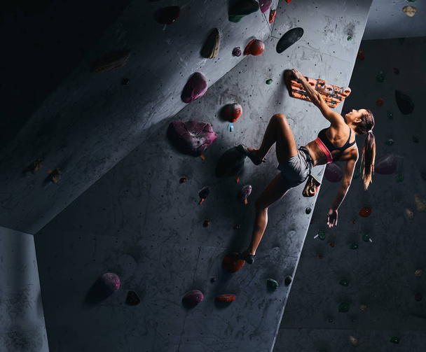 Young woman in shorts and sports bra exercising on a bouldering wall indoors. - Photo, Image