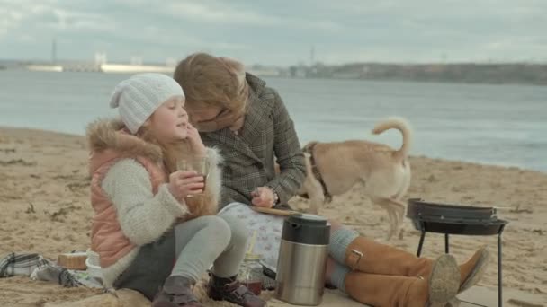 νεαρή γυναίκα με ένα παλτό με μια κοπέλα με τα σγουρά μαλλιά, μαμά και κόρη, κάθονται στην παραλία δίπλα στον ποταμό ωκεανό, έχοντας ένα πικ-νικ, πίνοντας ζεστό τσάι, κοπής λαχανικών, κοντά σε ένα σκυλί, κρύο - Πλάνα, βίντεο