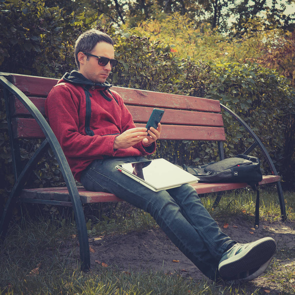 Fiatal csípő srác ellenőrzése bejelentés mobiltelefon, laptop számítógép, ül a parkban nyári nap távolság munka során. Jóképű férfi beszélgető online-ra smartphone alatt webinar-ra netbook - Fotó, kép