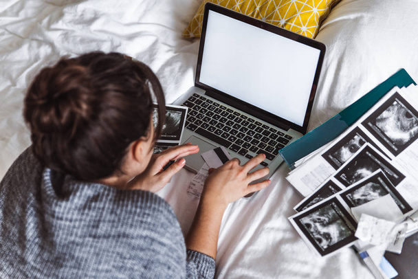 Amerikanische junge erwachsene Frau überprüft medizinische Ergebnisse mit Karte in der Hand. Laptop mit weißem Bildschirm auf dem Bett. Kopierraum. Gesundheitskonzept.. Krankenversicherung - Foto, Bild