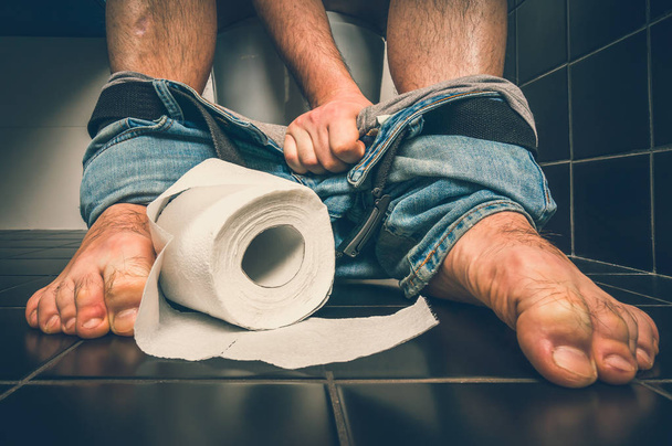 Man suffers from diarrhea is sitting on toilet bowl and toilet paper roll near his legs - diarrhea concept - retro style - Photo, Image