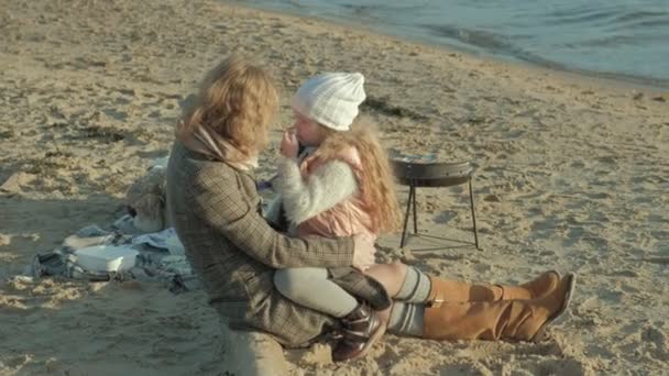 junge Frau im Mantel mit einem Mädchen mit lockigem Haar, Mutter mit Tochter, am Strand am Fluss, Meer, Picknick, Erholung, Wochenende, kaltes Wetter - Filmmaterial, Video