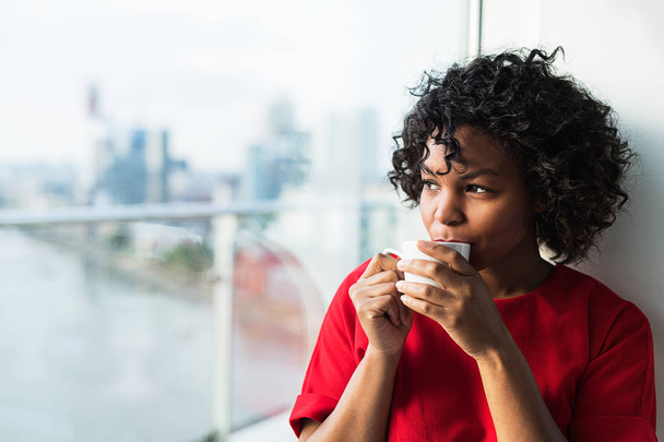 A close-up of a woman standing by the window drinking coffee. - Photo, Image