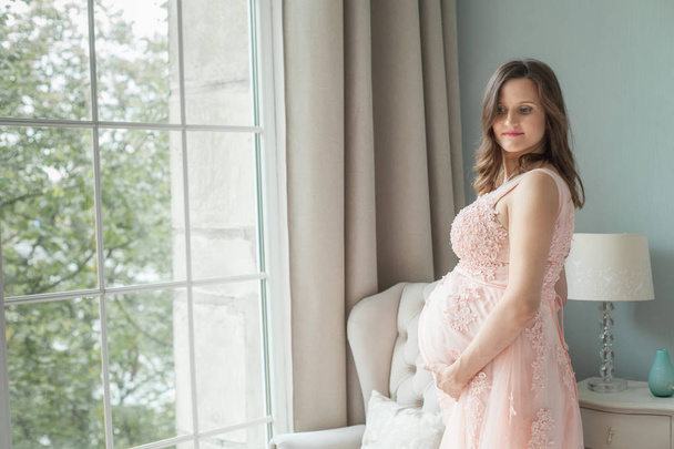 Pregnant woman in pink dress holds hands on belly at home. Pregnancy, maternity, preparation and expectation concept - Foto, afbeelding