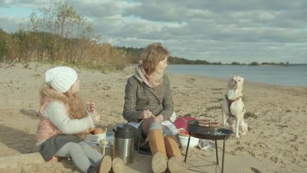 junge Frau im Mantel und ein Mädchen mit lockigem Haar, Mutter mit Tochter, sitzt am Strand am Fluss, Meer, hatte ein Picknick, Fleisch kochen auf dem Grill, neben einem Hund sitzen, kaltes Wetter - Filmmaterial, Video