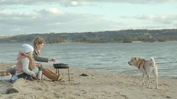 junge Frau im Mantel und Mädchen mit lockigem Haar, Mutter mit Tochter, sitzt am Strand am Fluss, Meer, picknickt, kocht Fleisch auf dem Grill, das Mädchen füttert den Hund, kaltes Wetter - Filmmaterial, Video
