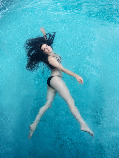 beautiful mature woman in the prime age with dark curly hair in a bikini is running floating weightless elegant happy floating in turquoise blue water in the pool. - Photo, Image