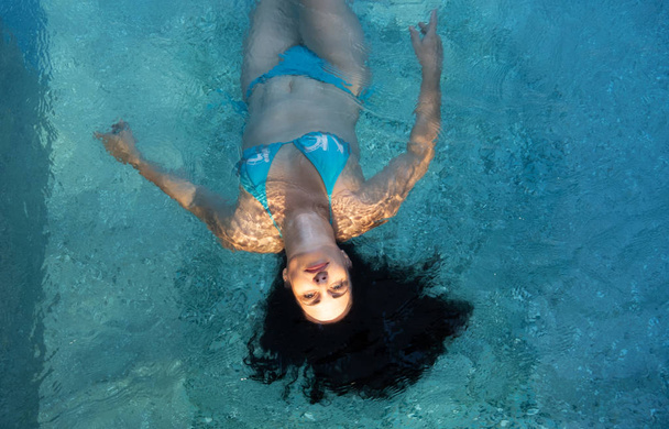 belle femme mature dans le meilleur âge avec des cheveux bouclés foncés en bikini bleu au soleil Lumière du soleil flottant en apesanteur élégamment heureux flottant dans l'eau turquoise bleue dans la piscine
 - Photo, image