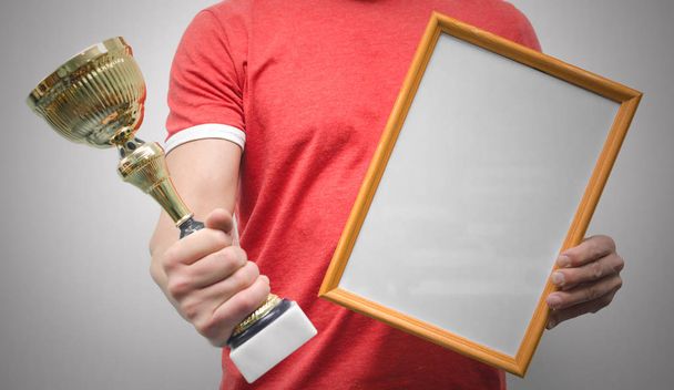 Man is holding a blank diploma or certificate frame with copy space and golden award trophy in the hands isolated on gray background. - Photo, Image