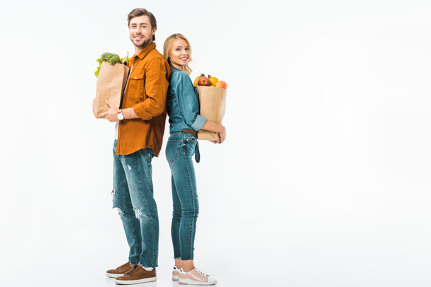glückliches Paar mit Einkaufstaschen voller Produkte, die Rücken an Rücken isoliert auf weiß stehen - Foto, Bild