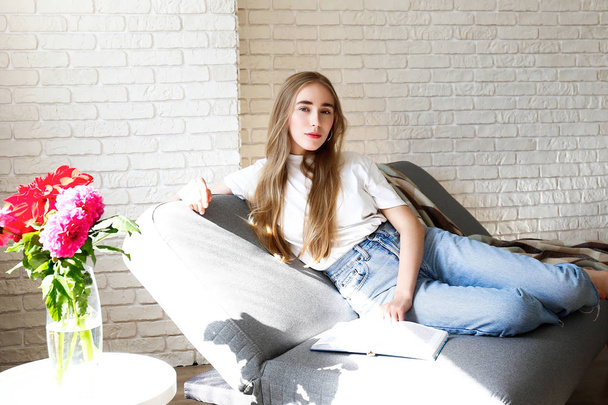 Young beautiful woman wearing white t-shirt on grey textile sofa at home. Attractive slim female in domestic situation, resting on couch in her lofty apartment. Background, copy space, close up. - Foto, Imagem