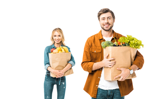 guapo sonriente hombre sosteniendo bolsa de compras con productos mientras su novia de pie detrás aislado en blanco
 - Foto, Imagen