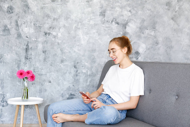 Young beautiful woman wearing white t-shirt on grey textile sofa at home. Attractive slim female in domestic situation, resting on couch in her lofty apartment. Background, copy space, close up. - Foto, imagen