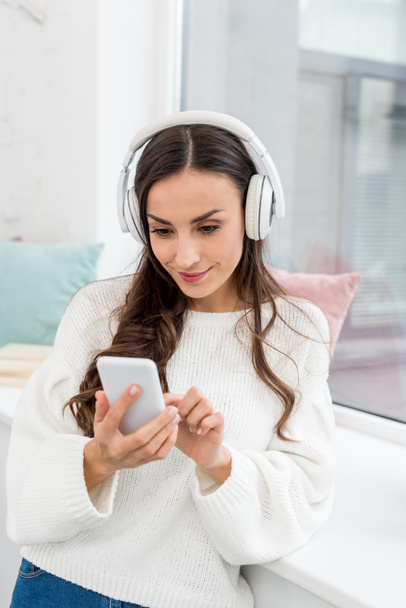 smiling young woman listening music with smartphone and wireless headphones ner window - Photo, Image