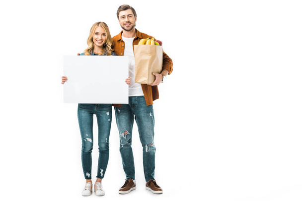 bonita mujer mostrando banner en blanco mientras su novio de pie con productos en bolsa de papel
  - Foto, Imagen