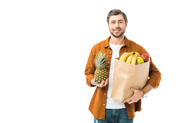 smiling man holding pineapple and paper bag full of products isolated on white - Foto, afbeelding