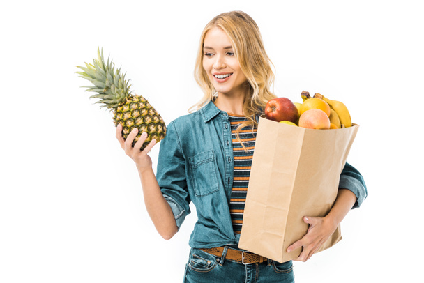 mulher alegre olhando para abacaxi e segurando saco de papel com produtos isolados em branco
 - Foto, Imagem