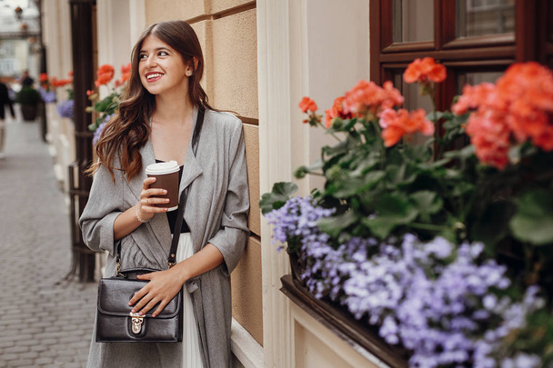 Stylish hipster girl with beautiful hair holding coffee cup and smiling in city street . Gorgeous young woman with paper cup of coffee to-go enjoying time and relaxing in morning - Photo, Image