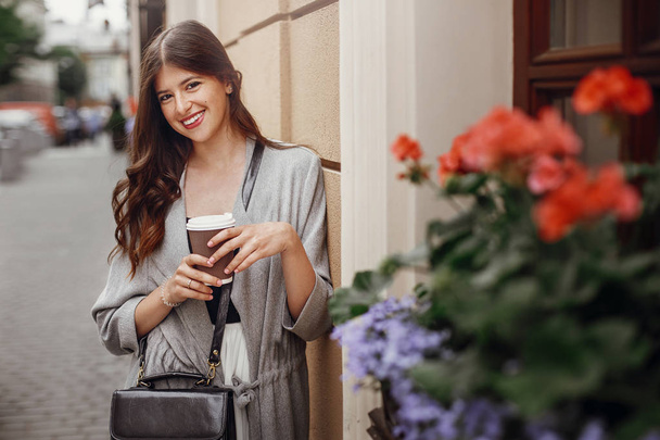 Gorgeous young woman with cup of coffee smiling and posing in city street. Stylish hipster girl with beautiful hair and perfect white smile enjoying time in the city. Copy space - Photo, Image