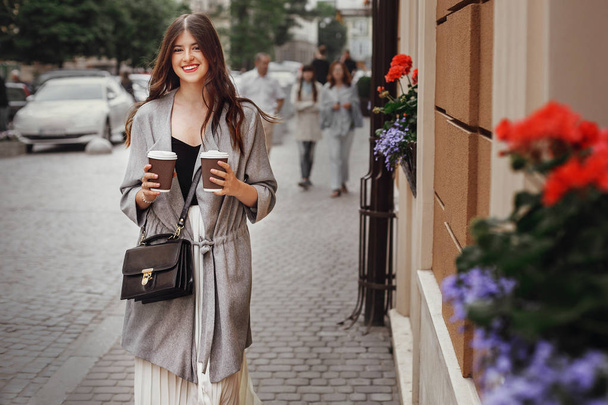 Stylish hipster girl with beautiful hair and smile holding two coffee cups and walking in city street . Gorgeous happy young woman with paper cups of coffee to-go enjoying time and smiling - Photo, Image