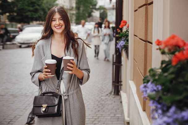 笑顔で街を歩いて、コーヒーを 2 杯と豪華な若い女性。美しい髪と完璧な笑顔の街で時間を楽しむスタイリッシュな幸せな内気な少女。コピー スペース - 写真・画像
