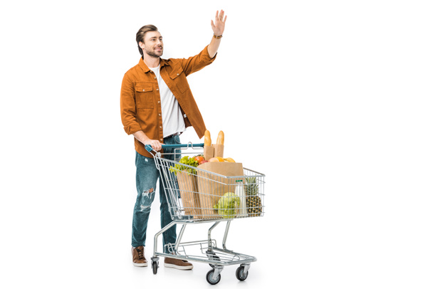 Ein lächelnder Mann winkt mit der Hand und steht neben einem Einkaufswagen mit Produkten in Papiertüten auf weißem Grund. - Foto, Bild