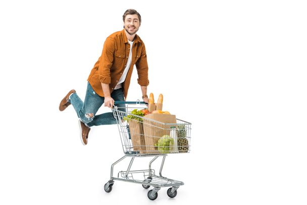 hombre positivo saltando y llevando carrito de compras con productos en bolsas de papel aisladas en blanco
 - Foto, Imagen