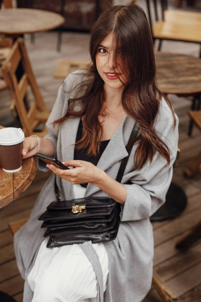 Gorgeous νεαρή γυναίκα κρατώντας τηλέφωνο και χαμογελαστός, με καφέ ξύλινα ρουστίκ τραπέζι στην βεράντα στο δρόμο της πόλης. Κομψό hipster κορίτσι με τα όμορφα μαλλιά και τέλειο χαμόγελο, περιήγηση στο internet - Φωτογραφία, εικόνα