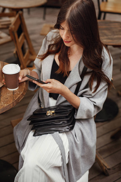 Upea nuori nainen, jolla on puhelin ja hymyilevä, kuppi kahvia puisella maalaispöydällä terassilla kaupungin kadulla. Tyylikäs hipster tyttö kauniit hiukset ja täydellinen hymy selailu internet
 - Valokuva, kuva