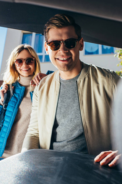 smiling man putting luggage into car while girlfriend standing behind on street - Foto, Imagen