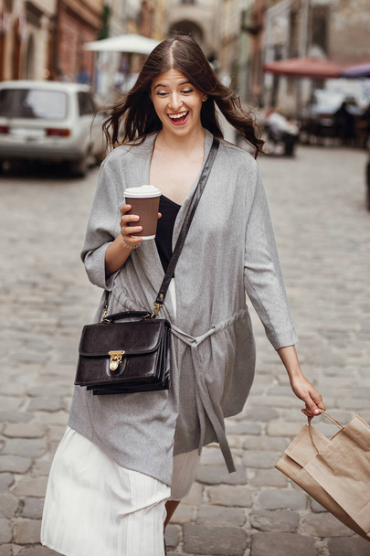 Hermosa joven mujer sosteniendo una taza de café y una bolsa, sonriendo y caminando por la calle de la ciudad. Elegante chica hipster feliz con hermoso cabello y emociones divertidas disfrutando del tiempo y las compras
 - Foto, imagen