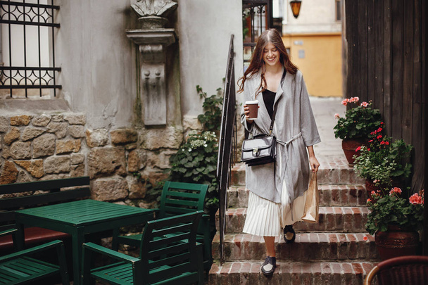 Gorgeous young woman holding cup of coffee and bag, smiling and walking in city street. Stylish happy hipster girl with beautiful hair, enjoying time at cafe terrace. Copy space - Zdjęcie, obraz
