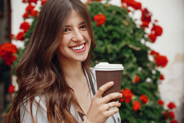 Stylish hipster girl with beautiful hair and perfect smile holding coffee cup  in city street on background of flowers. Gorgeous happy young woman enjoying time at cafe terrace. Copy space - Photo, Image